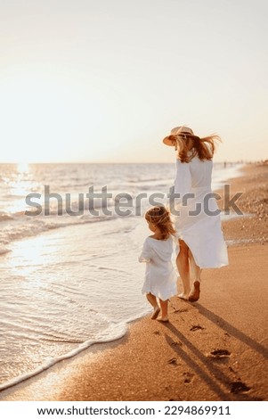 A little girl and a young mother in white dresses and a hat walk barefoot on the sand on the ocean at sunset. Wind blows hair