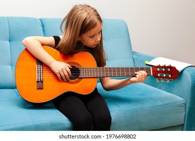 Little girl, young school age child playing guitar, focused kid practicing musical instrument sitting on the sofa at home, closeup. Talent, hobby, skills development concept, people lifestyle