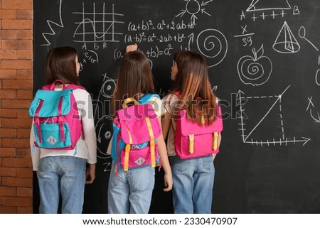 Little girls with backpacks near blackboard, back view