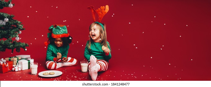 Little Girls Enjoying The Artificial Snow Flakes Falling Sitting Beside A Christmas Tree. Kids In Fancy Christmas Clothes Sitting Beside A Christmas Tree Eating Cookies With Milk.