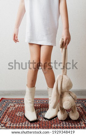 A little girl's feet in her mom's white boots. A little girl in her mom's white boots is holding a stuffed toy bunny by the ear.