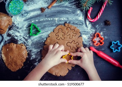 Little girl's hand making traditional Christmas cookies. Raw dough and cutters for the holiday cookies on a dark table. Preparing Christmas gingerbread cookies. Steps of making biscuits. Top view.