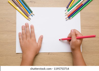 Little girl's hand painting on the white paper with pink pencil. Color pencils on the wooden desk. Drawing time. Top view. Empty place. - Powered by Shutterstock