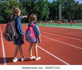 Little Girls School Children With Backpacks In The Stadium, Watching The Boys Play Football.