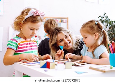 Little girls and smiling educator studying geometric figures, playing and folding cubes on desk sitting on floor in playroom. Interesting lesson for kindergartners developing logic and intelligence.