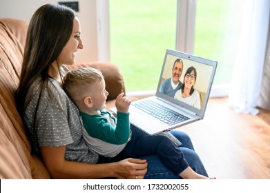 Little grandson is talking via video call on laptop with a grandparents. A young mother with a baby son sit on a sofa at home and have a video meeting, grandparent are on a laptop screen