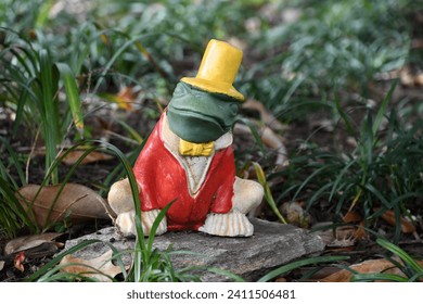 A little green frog wearing a yellow had and red blazer along with a bow tie. This little piece of garden decoration is super cute as he sits on a rock surrounded by a lush green background.