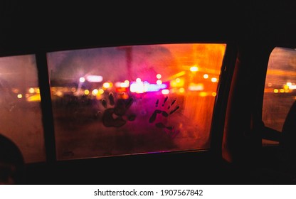 Little Kid’s Handprint On Wet Car Window Covered With Condensation In Night. Child’s Fingerprints On Glass Against Lights In Darkness.