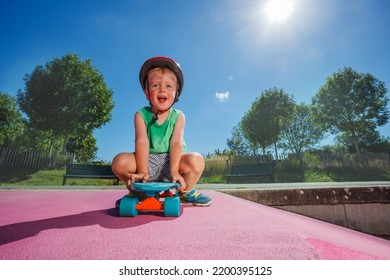 Little Handsome Smiling Boy Sit On The Skate Playing At Skatepark Wearing Helmet On Sunny Summer Day