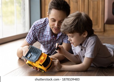 Little Handyman. Focused Son Kid Lying On Warm Wood Floor Fix Toy Car With Help Of Young Daddy. Junior Boy Listen To Father Advices Learn To Use Screwdriver Tool Imitating Dad Activity At Repair Work