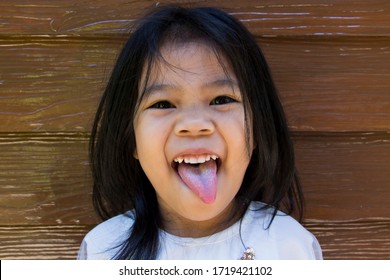 Little Happy Asian Girl Smiling With A Funny Face And Showing Her Tounge Looking At Camera On Wooden Background. Healthy Child, Family, Holiday Concept.