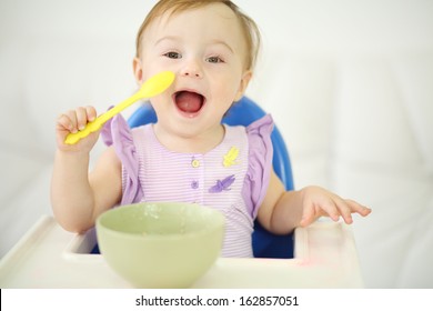 Little happy baby with spoon sits at highchair and eats porridge on plate. Shallow depth of field.