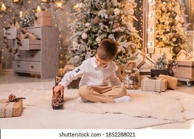 
Little Happy Boy Playing On The Floor With A Toy Vintage Steam Train, In The Living Room Against The Background Of The Christmas Tree. Time Of Miracles And Fulfillment Of Desires. Merry Christmas.