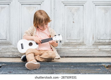 Little happy boy plays his guitar or ukulele, sitting by the wooden door outdoors