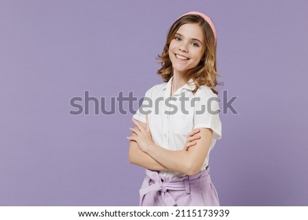 Little happy caucasian blonde kid girl 12-13 years old in white short sleeve shirt hold hands crossed folded isolated on purple color background children studio portrait. Childhood lifestyle concept