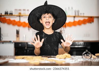 Little happy cheerful boy in the kitchen in a wizard's hat is preparing for Halloween to bake cookies, dirty all in flour, showing two palms. Play with food, bake dessert, prepare for Halloween party.