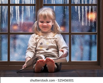 Little Happy Child Reading A Book. A Large Picture Window. Outside The Window, Snow Falls, Winter. The House Is Warm, The Girl Barefoot