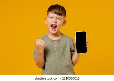Little Happy Fun Boy 6-7 Years Old In Green T-shirt Hold Use Mobile Cell Phone With Blank Screen Workspace Area Do Winner Gesture Isolated On Plain Yellow Background Mother's Day Love Family Concept