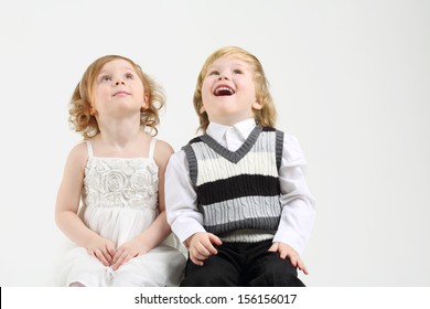 Little Happy Girl And Boy Sit On White Big Cube And Look Up On White Background.