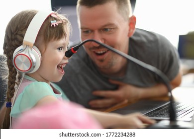 Little Happy Girl In Headphones Looks With Enthusiasm At Monitor Next To Man. Hanging Out Dad And Daughter