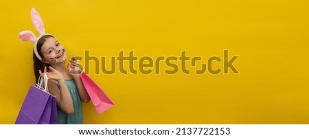 A little happy girl with long dark hair in rabbit ears on her head holds colorful Easter shopping bags on a yellow background, Happy Easter concept, Easter shopping, banner