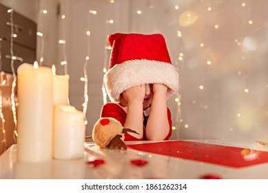 Little Happy Girl In A Red Dress And Hat Writes A Letter To Santa Claus. The Child Makes A Wish For Christmas. The Baby Does Not Know What To Make A Gift, She Covered Her Face With Hands. Copy Space