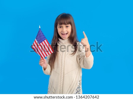 Little happy girl with USA flag shows one finger points upwards on a blue background.