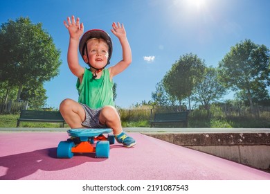 Little Happy Handsome Boy Sit On The Skate Playing At Skatepark Wearing Helmet Lift Hands On Sunny Summer Day