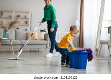 Little Helper. Adorable Toddler Boy Helping Mom Cleaning At Home, Kid Getting Rag Out Of Bucket While Mom Mopping Floor On Background, Cleaning Together In Living Room