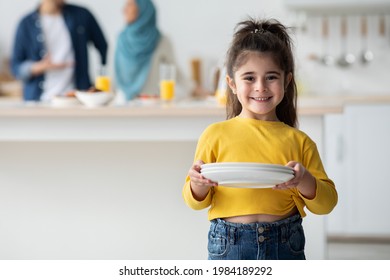 Little Helper. Portrait Of Cute Arab Girl Holding Plates In Kitchen And Smiling At Camera, Child Helping Parents To Prepare Table For Lunch At Home, Her Islamic Mom And Dad Chatting On Background