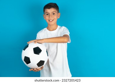 Little hispanic boy wearing white T-shirt holding a football ball gesturing with hands showing big and large size sign, measure symbol. Smiling looking at the camera.