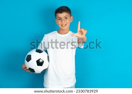 Little hispanic boy wearing white T-shirt holding a football ball making fun of people with fingers on forehead doing loser gesture mocking and insulting.