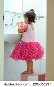 Little Independent Toddler Girl In Dress Standing On Kids Wood Chair In Bathroom. Cute Kid Washing Hands With Water And Soap. Curly Sweet Baby Play In Water. Healthy, Child's Hygiene Concept.