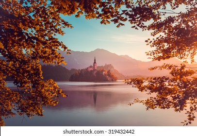 Little Island with Catholic Church in Bled Lake, Slovenia  at Sunrise with Castle and Mountains in Background. Autumn Filter. Tree Leaves Border. Natural Frame.