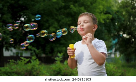 Little joyful boy in t-shirt blows soap bubbles playing in summer city park. Cheerful boy blows bubbles in park among trees on vacation. Schoolboy spends time blowing soap bubbles slow motion - Powered by Shutterstock