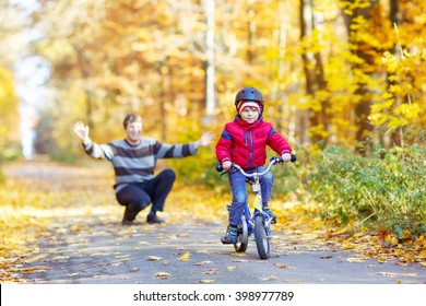 Little Kid Boy Of 3 Years And His Father In Autumn Forest With A Bicycle. Dad Teaching His Son. Man Happy About Success. Child With Helmet. Safety, Sports, Leisure With Kids Concept.