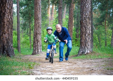 Little Kid Boy Of 4 Years And His Grand Father In Summer Forest With A Bicycle. Grandpa Teaching His Boy. Man Happy About Success. Child With Helmet. Safety, Sports, Leisure With Kids Concept.