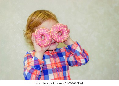 A little kid boy dabbles and plays with two fresh donuts before eating. A child holds donuts near his eyes and looks through the holes like through glasses.