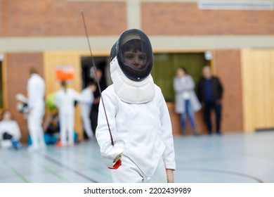 Little Kid Boy Fencing On A Fence Competition. Child In White Fencer Uniform With Mask And Sabre. Active Kid Training With Teacher And Children. Healthy Sports And Leisure.