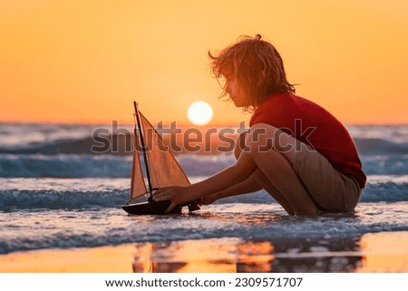 Little kid boy play with toy boat in the sea waves at the beach during summer vacation. Dream on travel. Sea dream. Dreamy kids face. Daydreamer child portrait close up. Dreams and imagination.