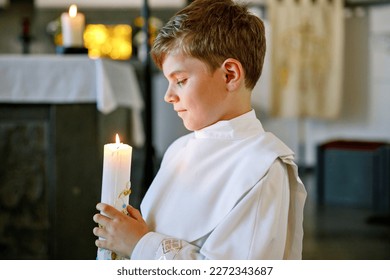 Little kid boy receiving his first holy communion. Happy child holding Christening candle. Tradition in catholic curch. Kid in a white traditional gown in a church near altar.