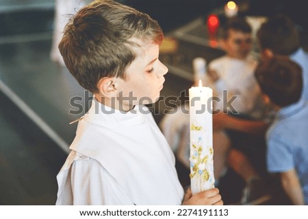 Little kid boy receiving his first holy communion. Happy child holding Christening candle. Tradition in catholic curch. Kid in a white traditional gown in a church near altar.