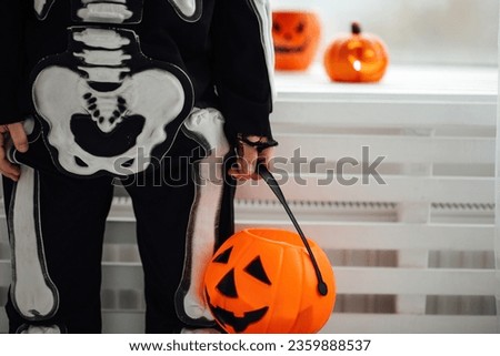 Little kid boy in skeleton costume with pumpkin candy bucket playing at home in hallows eve. Child is ready for trick or treat and scary gesture with jack lantern cup celebrates halloween