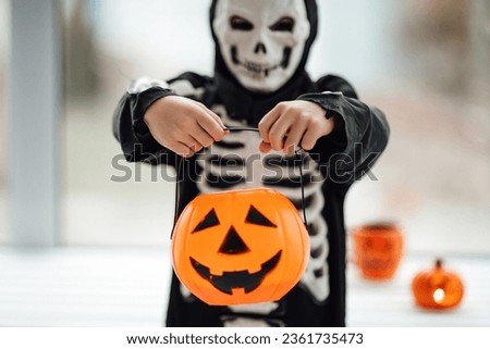 Little kid boy in skeleton costume with pumpkin candy bucket playing at home in hallows eve. Child is ready for trick or treat and scary gesture with jack lantern cup celebrates halloween