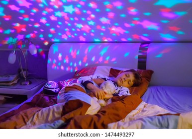 Little kid boy sleeping in bed with colorful lamp. School child dreaming and holding plush toy. Kid angry of darkness. Night light changing colors, stars and moon and playing music.