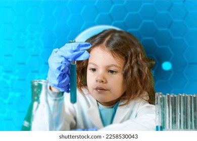 Little kid doing experiments with blue liquid. Cute child wants to become a doctor. Pretty child in white medical robe holds glass flasks with blue liquid on indigo background with hospital symbol. - Shutterstock ID 2258530043