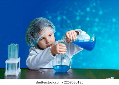 Little kid doing experiments with blue liquid. Cute child wants to become a doctor. Pretty child in white medical robe holds glass flasks with blue liquid on indigo background with hospital symbol. - Shutterstock ID 2258530135