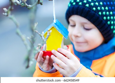 Little Kid Feeding Birds In Winter. Child Hanging Colorful Selfmade Bird House On Tree On Frosty Cold Day. Toddler In Colorful Wam Clothes. Selective Focus On Hands And Feeder With Seeds