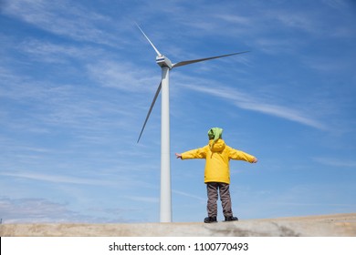 Little Kid In Front Of Windmill. Playful Child Looks At The Wind Turbine