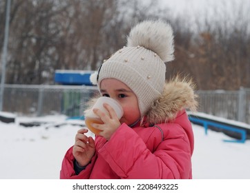 Little Kid Girl With A Cup Of Warm Cocoa Outdoors For Christmas. Child On A Walk In The City Park.Family Winter Vacation. An Alternative To Computer Games. A Girl In A Pink Jacket Drinks Hot Tea In A 